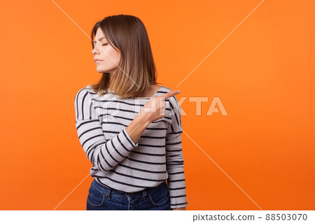 Get out. Portrait of resentful woman with brown hair in long sleeve shirt standing with closed eyes and offended face, pointing sideways showing exit. indoor studio shot isolated on orange background Get out. Portrait of resentful woman with brown hair in long sleeve shirt standing with closed eyes and offended face, pointing sideways showing exit. indoor studio shot isolated on orange background 88503070