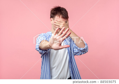 Don't want to look at this. Portrait of brown-haired man in casual striped shirt covering eyes with hand and showing stop gesture, refusing to watch. indoor studio shot isolated on pink background Don't want to look at this. Portrait of brown-haired man in casual striped shirt covering eyes with hand and showing stop gesture, refusing to watch. indoor studio shot isolated on pink background 88503075