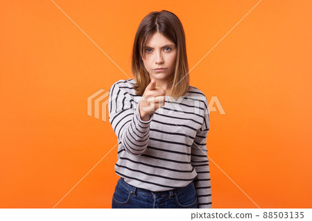 Hey you. Portrait of suspicious disgruntled young woman with brown hair in long sleeve striped shirt standing, pointing finger at camera and blaming. indoor studio shot isolated on orange background 88503135