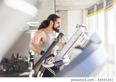 Closeup side view portrait of young adult sport man with long curly hair training alone. Handsome male doing exersices in a gym on elliptical machine and looking at window. Indoor, fitness concept 88503201