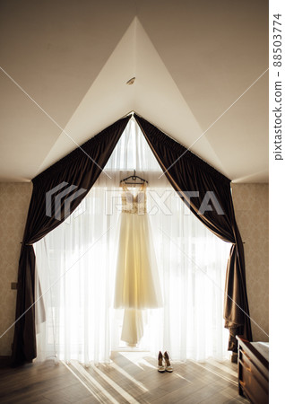 An elegant wedding dress hangs by a curtained window. Nearby are women's shoes An elegant wedding dress hangs by a curtained window. Nearby are women's shoes 88503774
