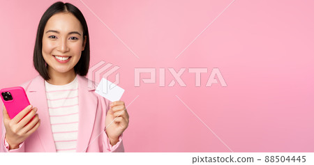 Image of smiling happy asian businesswoman showing credit card, paying online on smartphone application, order with mobile phon, standing against pink background Image of smiling happy asian businesswoman showing credit card, paying online on smartphone application, order with mobile phon, standing against pink background 88504445