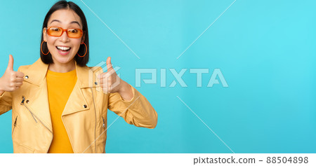 Enthusiastic korean girl looks excited and shows thumbs up, approves smth awesome, excellent choice and quality, stands over blue background 88504898