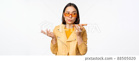 Image of happy asian girl talking on speakerphone, recording, translating her voice with mobile phone app, talking in smartphone dynamic, standing over white background 88505020