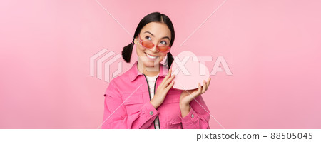 Image of stylish asian girlfriend in sunglasses, guessing whats inside gift box, heart-shaped present, standing over pink background Image of stylish asian girlfriend in sunglasses, guessing whats inside gift box, heart-shaped present, standing over pink background 88505045