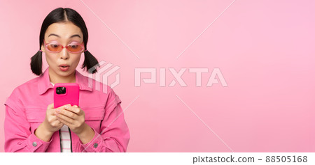 Portrait of asian girl in sunglasses using smartphone. Woman looking at mobile phone, browsing in app, standing over pink background 88505168