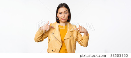 Image of unamused asian girl pointing at herself, looking with disbelief skeptical at camera, posing against white background 88505560