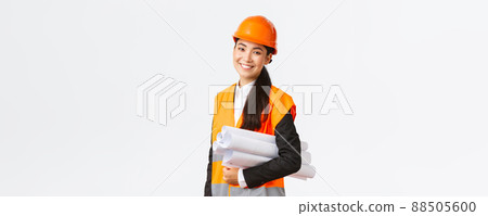 Smiling successful asian female architect, construction engineer in safety helmet, carry blueprints and smiling happy at camera, introduce building project, standing white background 88505600