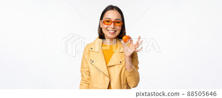 Beautiful asian girl in sunglasses showing tangerine and smiling, looking happy, posing in yellow against studio background 88505646