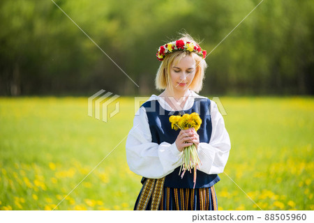 Young woman in national clothes wearing yellow dandelion wreath in spring field. Springtime Young woman in national clothes wearing yellow dandelion wreath in spring field. Springtime 88505960