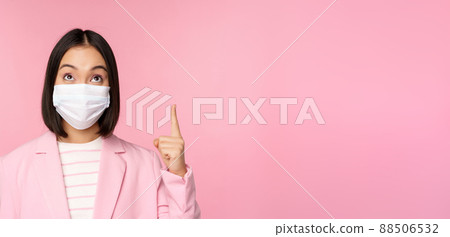 Close up portrait of businesswoman, asian lady in office suit and medical face mask, looking and pointing up, showing company logo or banner on top, pink background 88506532