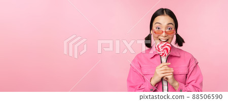 Silly and cute asian female model licking lolipop, eating candy sweet and smiling, looking excited, standing over pink background 88506590