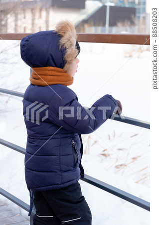 a pensive boy in a jacket and a hood stands by the fence in winter a pensive boy in a jacket and a hood stands by the fence in winter 88508203