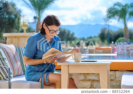Middle aged woman resting near outdoor pool reading book 88512264