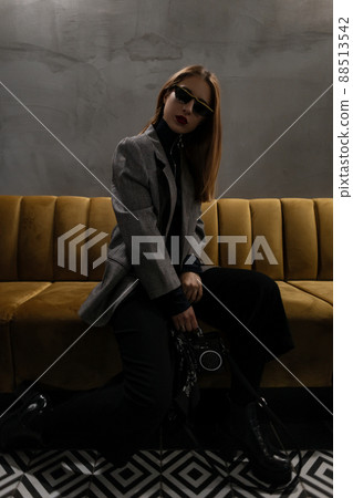 Young hipster woman in sunglasses with a pierced nose in a checkered jacket in retro style with a fashionable handbag-recorder posing sitting on a sofa in a cafe. Beautiful old fashioned punk girl. Young hipster woman in sunglasses with a pierced nose in a checkered jacket in retro style with a fashionable handbag-recorder posing sitting on a sofa in a cafe. Beautiful old fashioned punk girl. 88513542