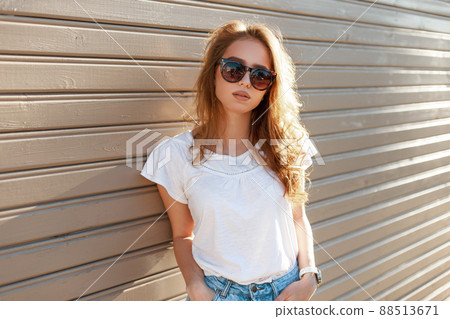 Young modern hipster woman in stylish sunglasses in a summer fashion white t-shirt in trendy jeans is standing near a wooden vintage wall on a sunny spring day. Cute girl resting outdoors. 88513671