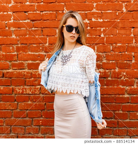 Beautiful sexy young hipster woman in a white lace blouse and denim jacket in black sunglasses in a beige skirt poses near a red brick wall. European modern girl. Beautiful sexy young hipster woman in a white lace blouse and denim jacket in black sunglasses in a beige skirt poses near a red brick wall. European modern girl. 88514134