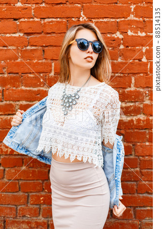 Attractive elegant young woman hipster in a black sunglasses in a summer denim jackets in a vintage white lace blouse in a skirt posing near the red brick wall outdoors. Fashionable modern girl. Attractive elegant young woman hipster in a black sunglasses in a summer denim jackets in a vintage white lace blouse in a skirt posing near the red brick wall outdoors. Fashionable modern girl. 88514135