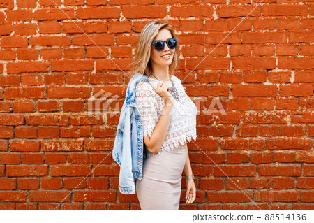 Charming happy young hipster woman with a beautiful smile in a white lace blouse in a denim jacket in black sunglasses in a beige skirt posing near a red brick wall. European joyful modern girl. Charming happy young hipster woman with a beautiful smile in a white lace blouse in a denim jacket in black sunglasses in a beige skirt posing near a red brick wall. European joyful modern girl. 88514136