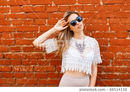 Happy attractive young hipster woman in sunglasses in a white lace blouse with a beautiful shiny necklace in a skirt posing near a vintage red brick wall in a summer day. European girl for a walk. Happy attractive young hipster woman in sunglasses in a white lace blouse with a beautiful shiny necklace in a skirt posing near a vintage red brick wall in a summer day. European girl for a walk. 88514138
