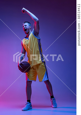 Studio shot of muscled man, basketball player training with ball isolated on purple background in neon light. Goals, sport, motion, activity concepts. 88514411