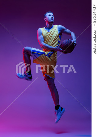 Studio shot of muscled man, basketball player training with ball isolated on purple background in neon light. Goals, sport, motion, activity concepts. 88514437