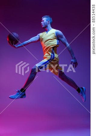 Studio shot of muscled man, basketball player training with ball isolated on purple background in neon light. Goals, sport, motion, activity concepts. 88514438