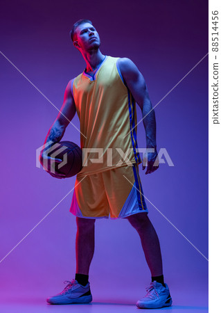 Studio shot of muscled man, basketball player standing with ball isolated on purple background in neon light. Goals, sport, motion, activity concepts. 88514456