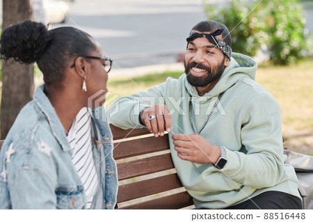 Smiling handsome bearded man in hoodie sitting on bench and telling funny story to black girl in city 88516448