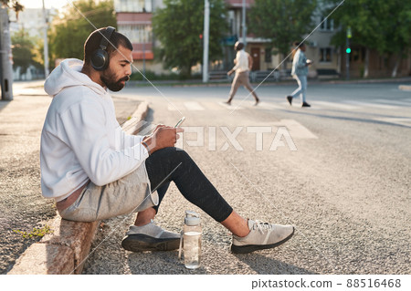 Athletic young middle eastern man in wireless headphones texting message on phone while relaxing on curb in city Athletic young middle eastern man in wireless headphones texting message on phone while relaxing on curb in city 88516468