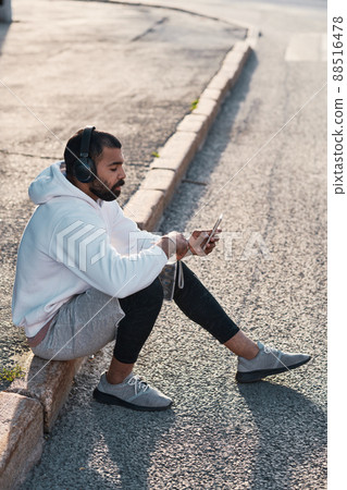 Calm young bearded man in white hoodie sitting on curb and using smartphone while enjoying music in headphones 88516478