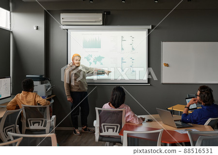 Young bearded man in hat and hoodie standing at projection screen and sharing statistics at meeting of staff Young bearded man in hat and hoodie standing at projection screen and sharing statistics at meeting of staff 88516531