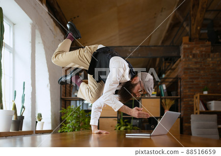 On line meeting. Young bearded man, office clerk having fun, doing yoga on wooden table in modern office at work time with gadgets. Concept of business, healthy lifestyle, sport, hobby 88516659