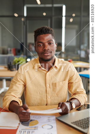 Portrait of content handsome young African-American marketing expert in yellow shirt sitting with papers in modern office Portrait of content handsome young African-American marketing expert in yellow shirt sitting with papers in modern office 88516920