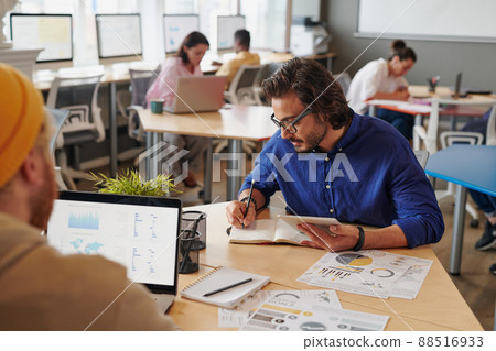 Concentrated young mixed race man in glasses sitting at table and making notes in organizer while planning new project 88516933