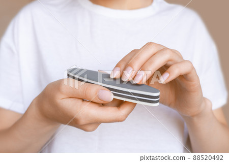 Female hands with natural manicure holding gray nail file. Woman in white holds emery board for smoothing and shaping the fingernails. Concept of self-care, beauty and professional tools usage Female hands with natural manicure holding gray nail file. Woman in white holds emery board for smoothing and shaping the fingernails. Concept of self-care, beauty and professional tools usage 88520492