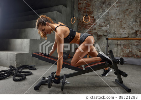 Close-up of side view of muscular athletic brunette woman who trains in gym with dumbbell in her hand on simulator. 88521288