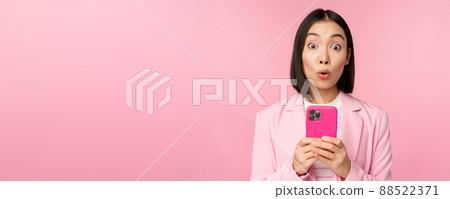 Portrait of asian businesswoman with surprised face, using smartphone app, wearing business suit. Korean girl with mobile phone and excited face expression, pink background 88522371