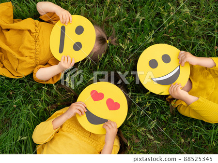 Three children lying on the grass are holding cardboard emoticons with different emotions in their hands Three children lying on the grass are holding cardboard emoticons with different emotions in their hands 88525345