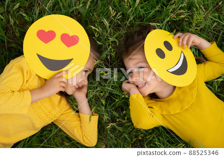 Two funny little girls are lying on the lawn covering part of their faces with emoticons and laughing merrily. World emoji Day. Anthropomorphic smile Face. 88525346