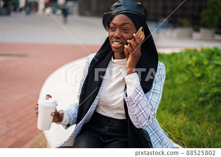 Joyful African American lady in trendy smart casual wear and Muslim headscarf sitting with cup of takeaway drink in urban park and talking on mobile phone 88525802