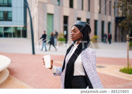 Side view of confident African American female in checkered blazer leather beret and black Muslim headscarf standing with cup of takeaway coffee in city 88525803