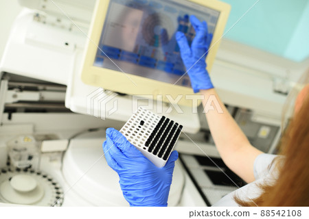 an an employee of a medical bacteriological laboratory holds a container with test tubes in his hands against the background of the biochemical analyzer. an an employee of a medical bacteriological laboratory holds a container with test tubes in his hands against the background of the biochemical analyzer. 88542108