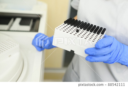 a close-up of an employee of a medical bacteriological laboratory holding a container with test tubes against the background of the biochemical analyzer. a close-up of an employee of a medical bacteriological laboratory holding a container with test tubes against the background of the biochemical analyzer. 88542111