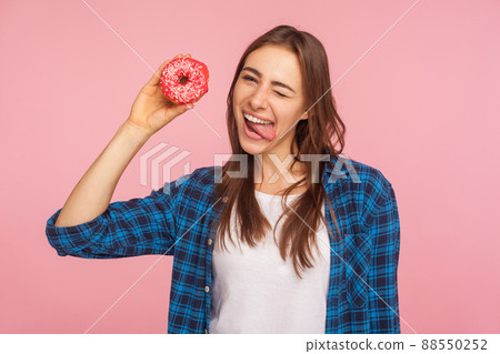 Confectionery and junk food. Portrait of joyous playful girl in checkered shirt sticking out tongue and holding doughnut, having fun with dessert. indoor studio shot isolated on pink background 88550252