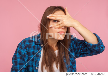 Portrait of nosy funny girl in checkered shirt peeking through fingers, looking with embarrassed inquisitive expression, watching secrets, spying rumors. indoor studio shot isolated on pink background 88550263