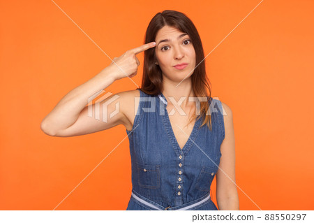 Are you kidding me. Positive brunette woman in denim dress holding finger near head temple and gesturing You are stupid idiot, looking displeased with crazy idea. indoor studio shot orange background Are you kidding me. Positive brunette woman in denim dress holding finger near head temple and gesturing You are stupid idiot, looking displeased with crazy idea. indoor studio shot orange background 88550297