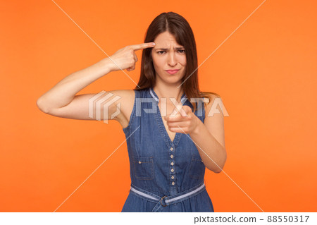 You are idiot. Positive brunette woman in denim dress holding finger near head temple and showing stupid gesture, pointing to camera with mocking expression. studio shot isolated on orange background 88550317