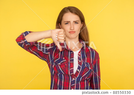 Dislike. Portrait of dissatisfied unhappy ginger girl in red shirt showing thumb down gesture and looking displeased, negative feedback, disapproval sign. studio shot isolated on yellow background Dislike. Portrait of dissatisfied unhappy ginger girl in red shirt showing thumb down gesture and looking displeased, negative feedback, disapproval sign. studio shot isolated on yellow background 88550330