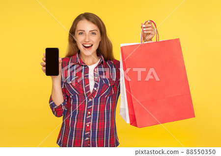 Amazed surprised ginger girl in casual style checkered shirt holding shopping bags and mobile phone, advertising online store, looking shocked. indoor studio shot isolated on yellow background Amazed surprised ginger girl in casual style checkered shirt holding shopping bags and mobile phone, advertising online store, looking shocked. indoor studio shot isolated on yellow background 88550350